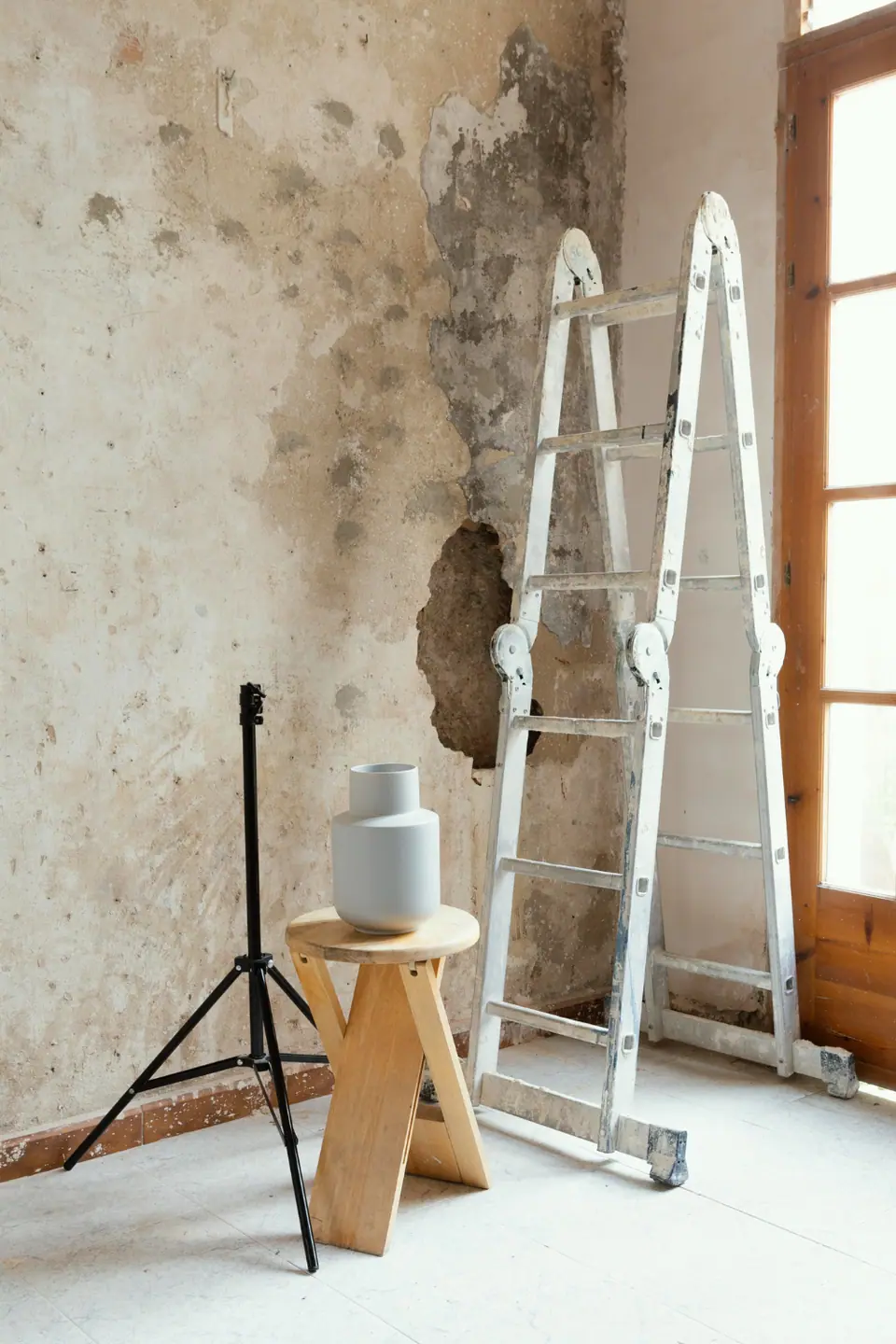 A metal ladder, wooden stool with stacked grey cylinders, and a black tripod stand are placed in a room with a damaged, peeling wall and a window.