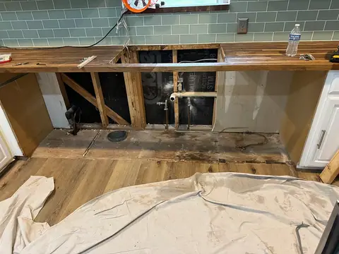 Kitchen renovation scene showing removed lower cabinets and appliances, exposed plumbing and wall framing, with tools and materials on a bare wood countertop and a drop cloth on the floor—highlighting potential DIY cleanup risks.