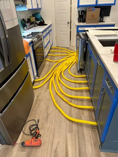 Yellow hoses are spread across a kitchen floor, running into cabinets and appliances; tools lie nearby, and blue tape covers the cabinet edges—evidence of water damage restoration in progress for concerned homeowners.