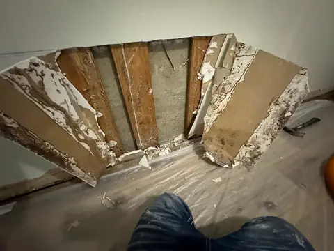 A section of drywall has been removed, exposing wooden studs and spot mold or water damage; plastic sheeting covers the floor to help contain this potential health hazard, while a person’s knees are visible in the foreground.
