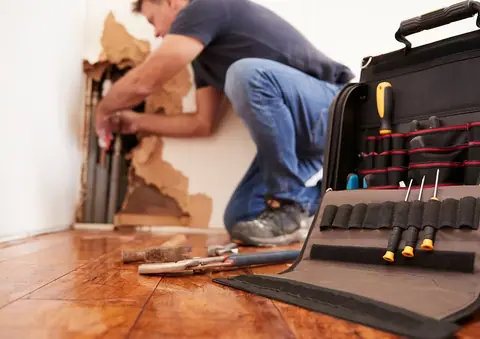 A person repairs plumbing inside a wall with exposed pipes, helping to prevent water damage and mold growth; tools and a tool bag are on the wooden floor nearby.