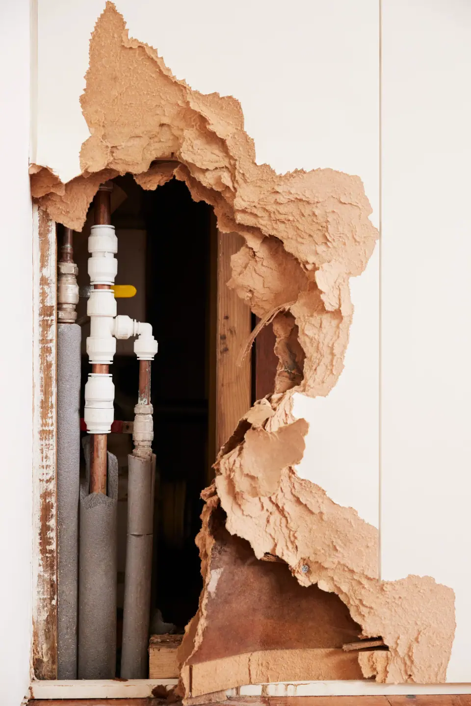 A large hole in a wall reveals exposed plumbing pipes and insulation, likely caused by a burst pipe in Winnsboro, TX, with crumbling drywall edges surrounding the opening.